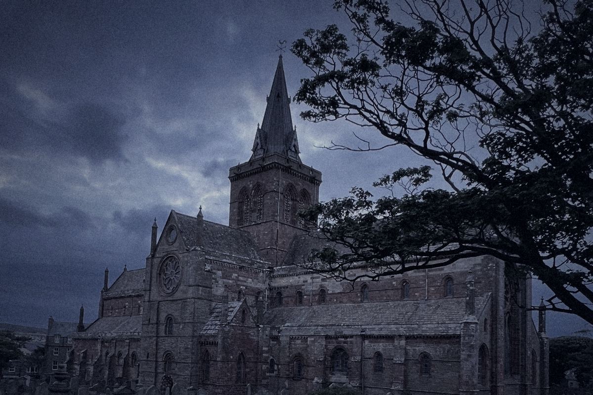 The tower and spire of St Magnus Cathedral in Kirkwall at nightfall, behind a windswept tree.